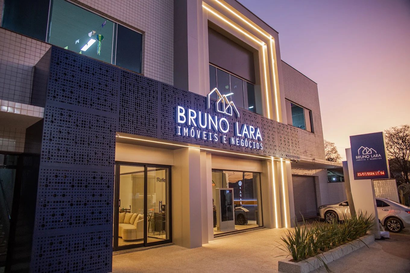 Fachada da Bruno Lara Imóveis e Negócios ao entardecer, com painel perfurado azul, letreiro iluminado e frisos de luz
